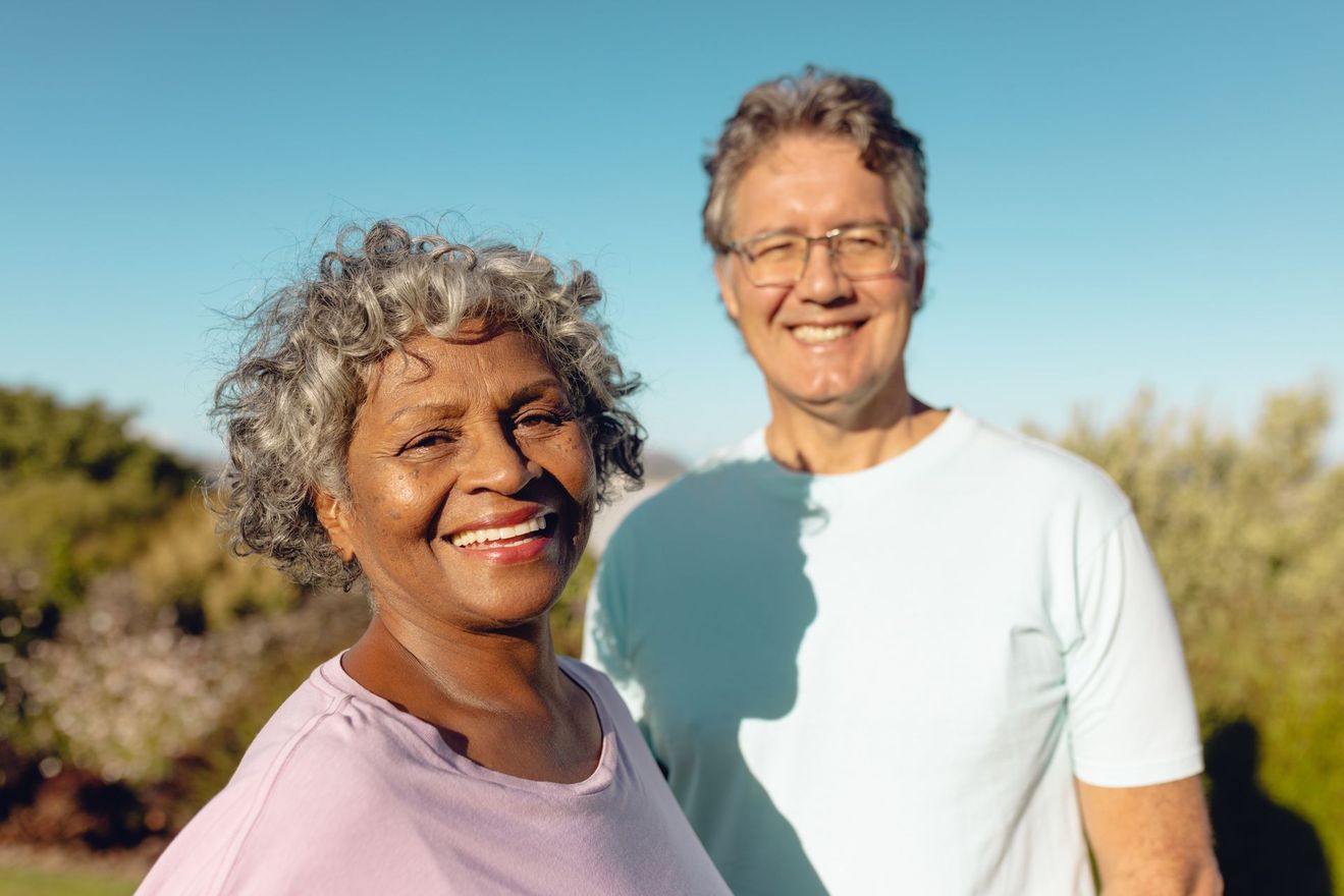 Two smiling people standing outdoors in a sunny field with trees in the background