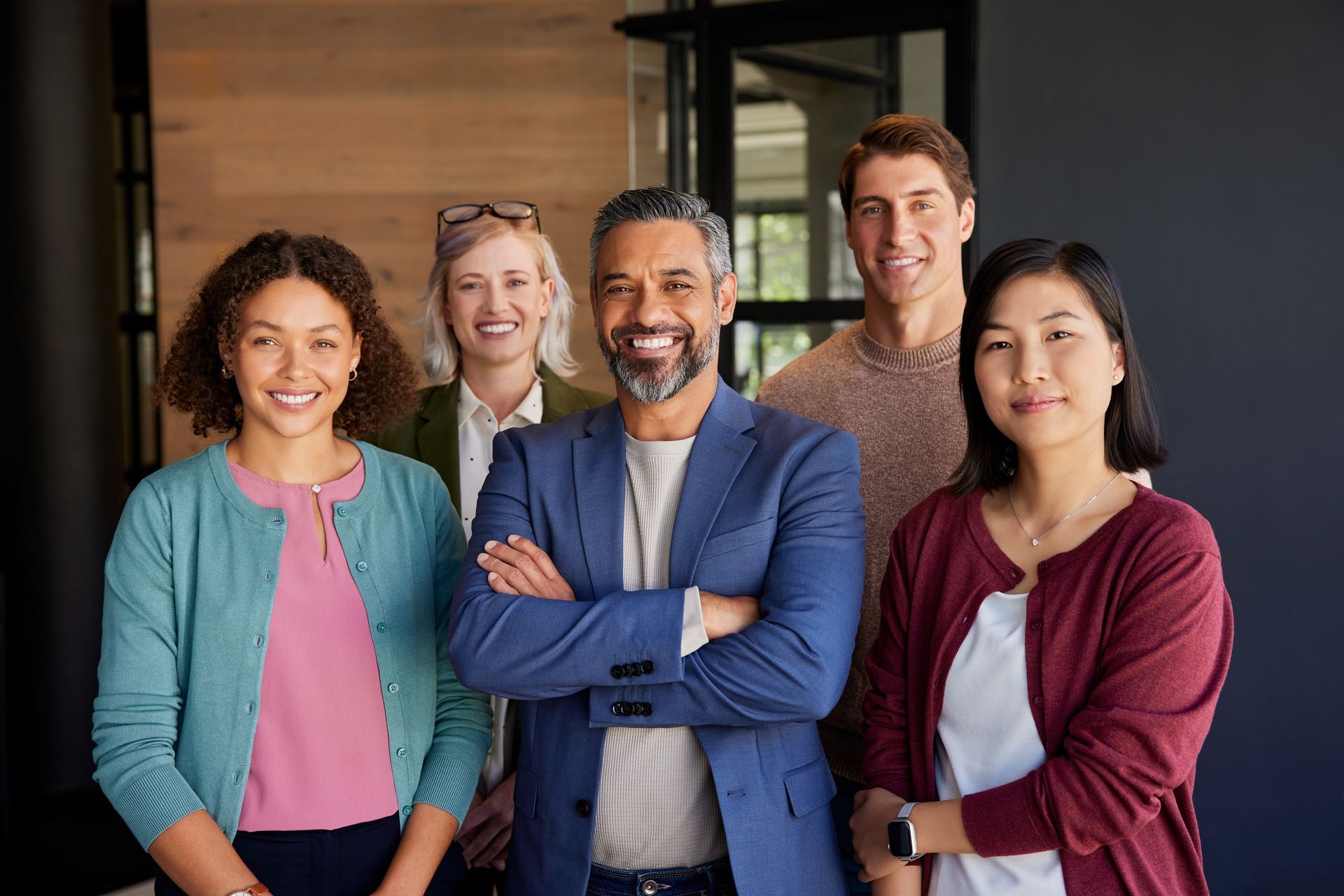 Five smiling coworkers stand indoors with arms crossed in a bright office hallway.