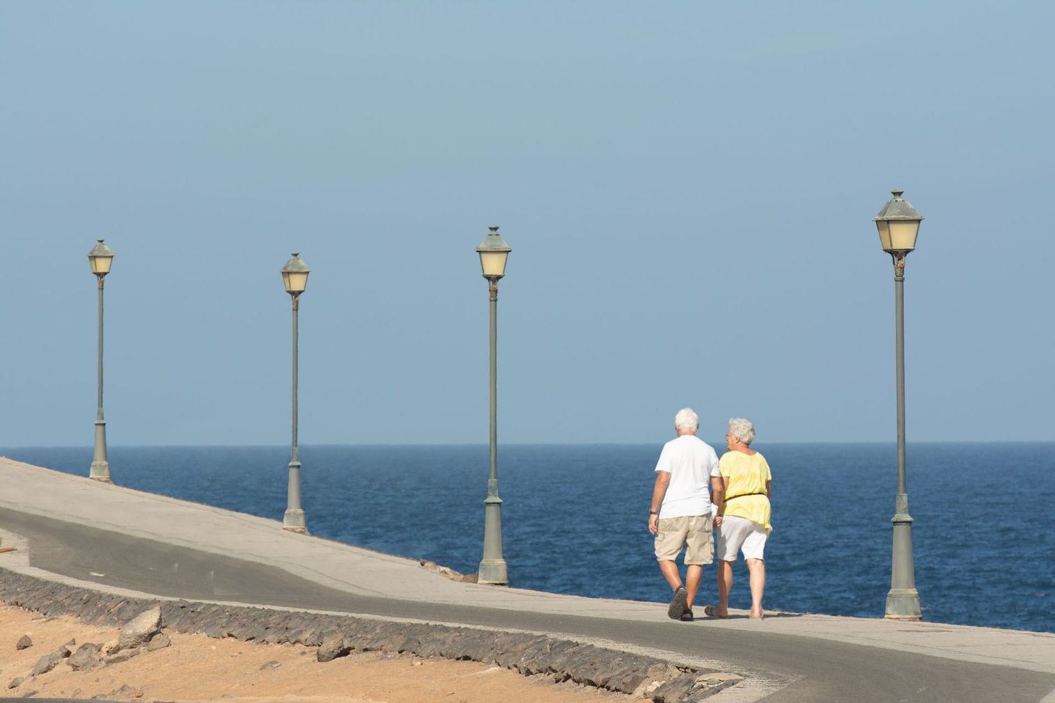 Two people walking along a seaside promenade beside lampposts and the blue ocean