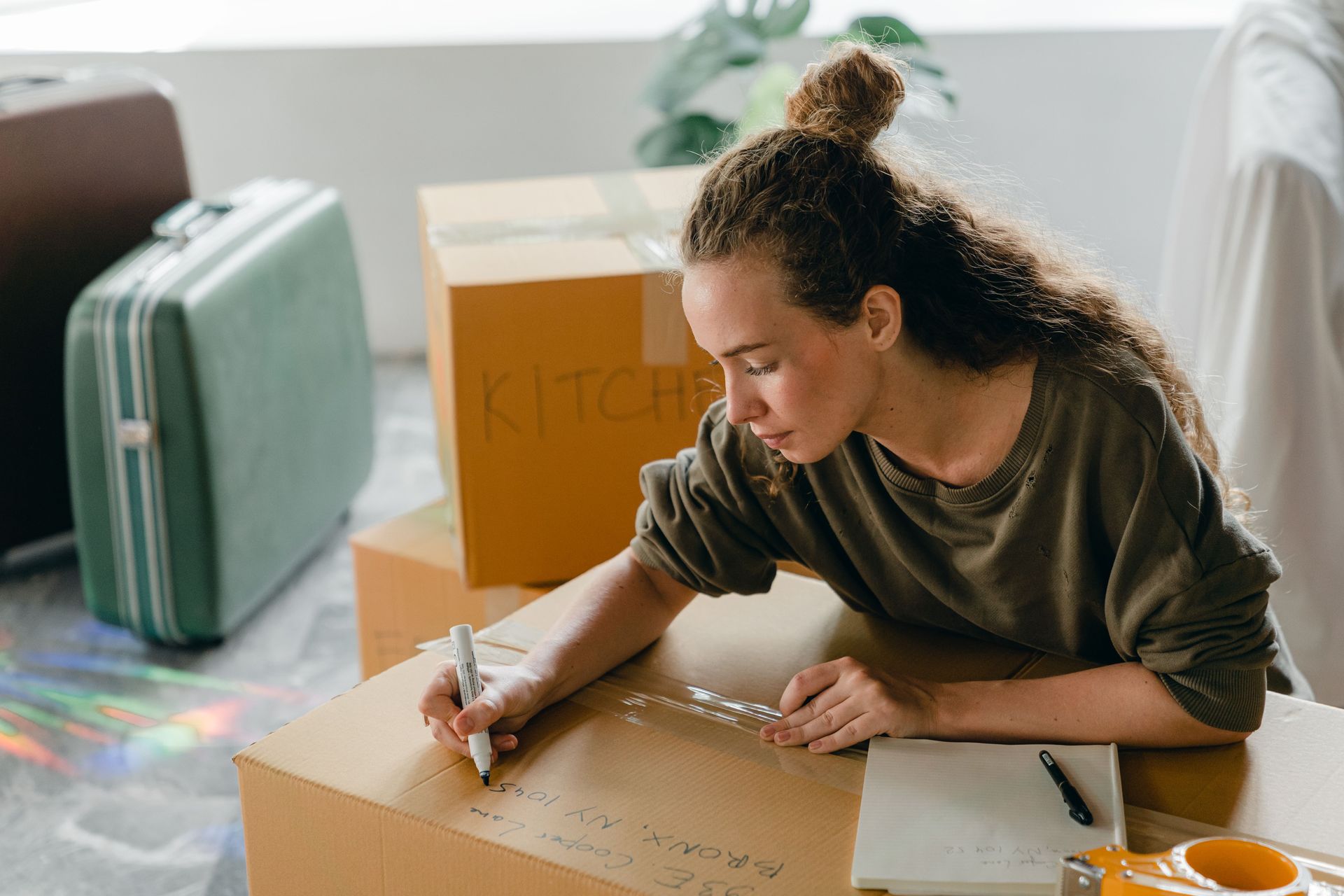 Woman kneeling on the floor, labeling a cardboard box with a marker beside a suitcase and packing materials.
