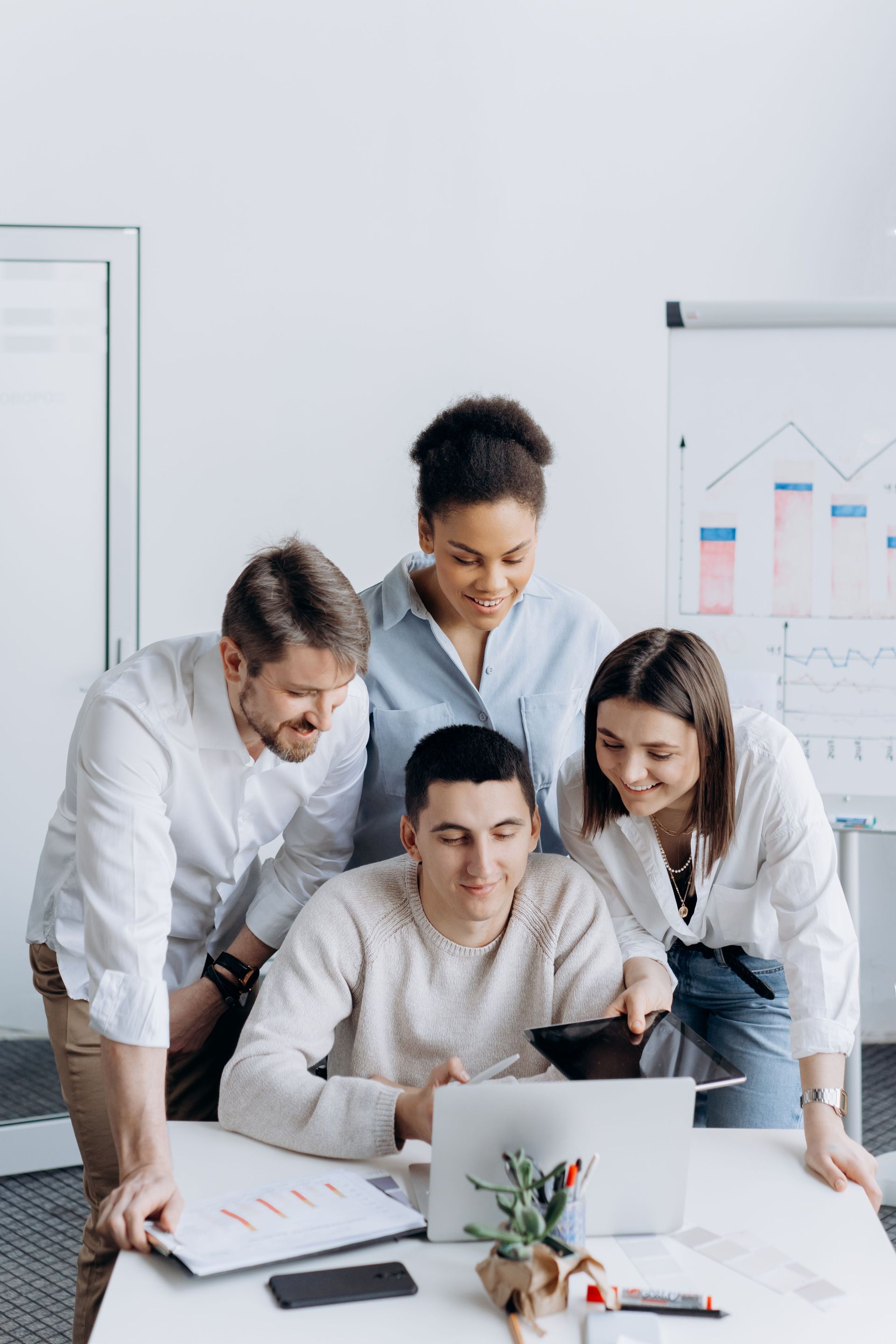 Four coworkers collaborate around a laptop at a desk in a bright office, with charts in the background.