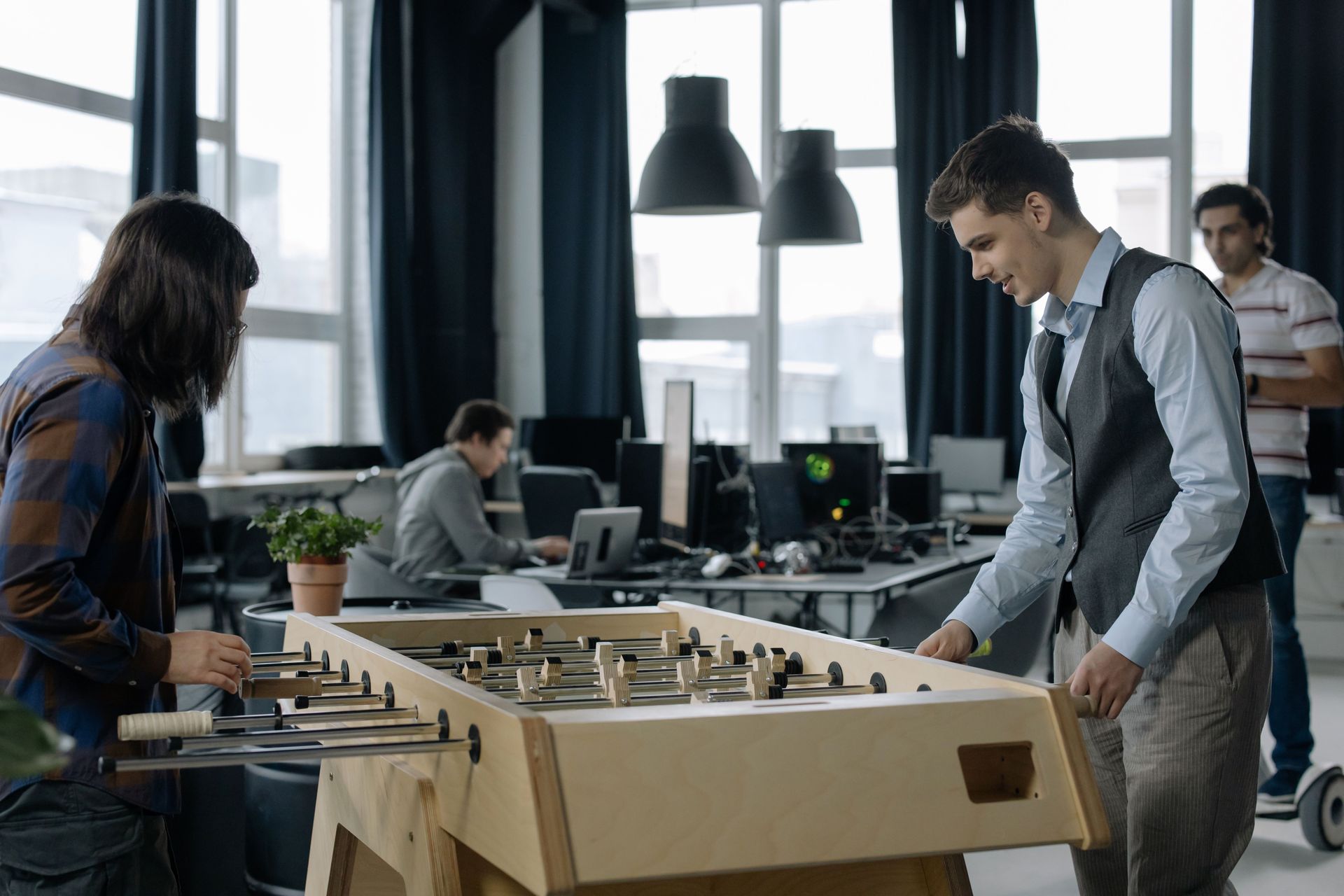 People playing foosball in a modern office lounge, with coworkers nearby at desks.