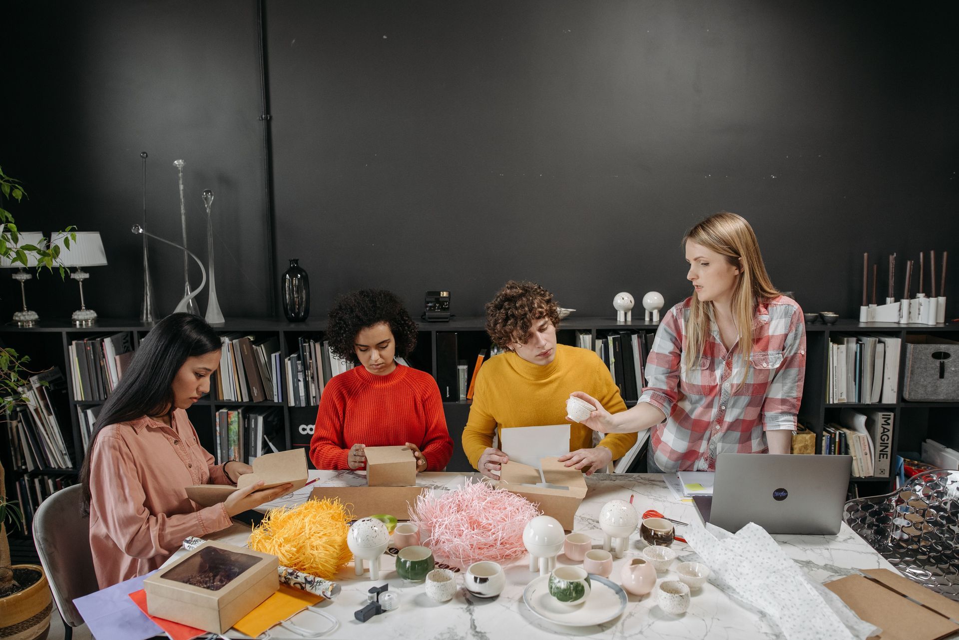 Four people collaborate at a table with flowers, notebooks, and a laptop in a dark studio office.