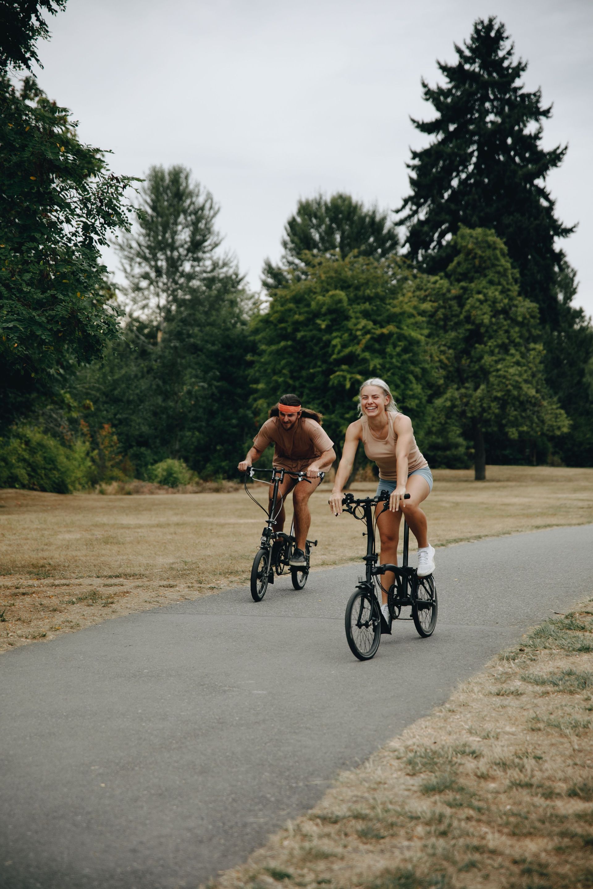 Two cyclists riding on a paved path beside dry grass and tall trees
