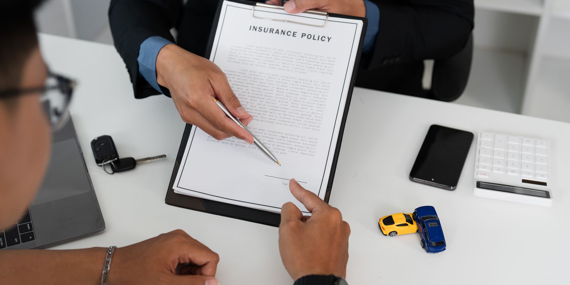 An agent pointing to a clipboard insurance policy while a client signs, toy cars and a phone nearby An agent pointing to a clipboard insurance policy while a client signs, toy cars and a phone nearby