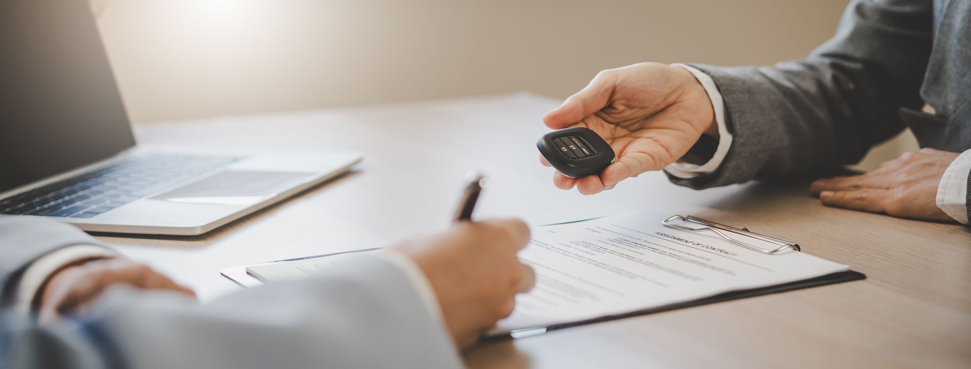 A person handing car keys to a customer signing insurance paperwork on a clipboard at an office desk