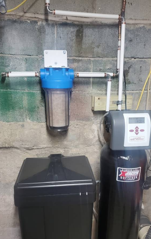 Water filtration system with a blue filter housing and black tank against a concrete wall.