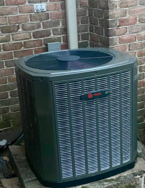 Dark gray Trane air conditioning unit against a brick wall, with a white pipe and electrical box nearby.