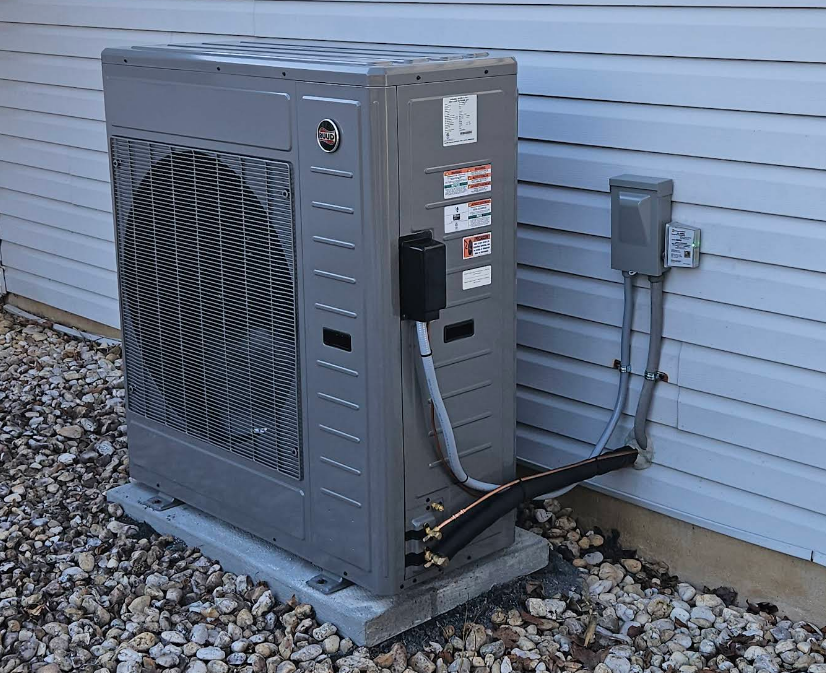 Gray heat pump unit on a concrete pad, gravel ground, next to a house with gray siding.