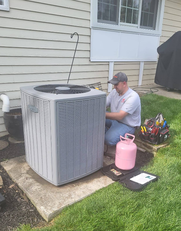 technician kneeling by an air conditioning unit; pink refrigerant tank, tools on a mat, and a white house in the background.