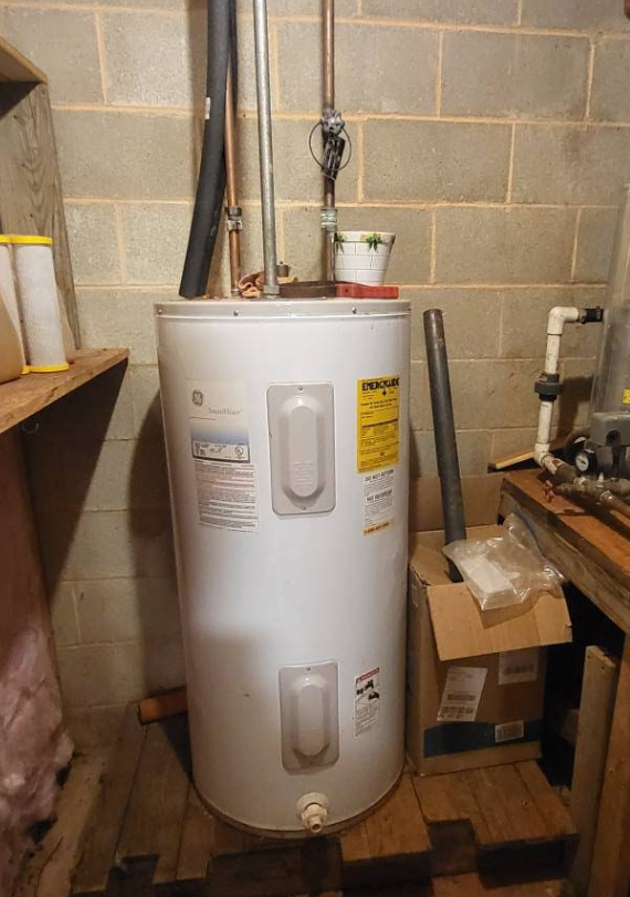 Water heater in a basement. White tank with insulation, various pipes, and wooden structures in the background.