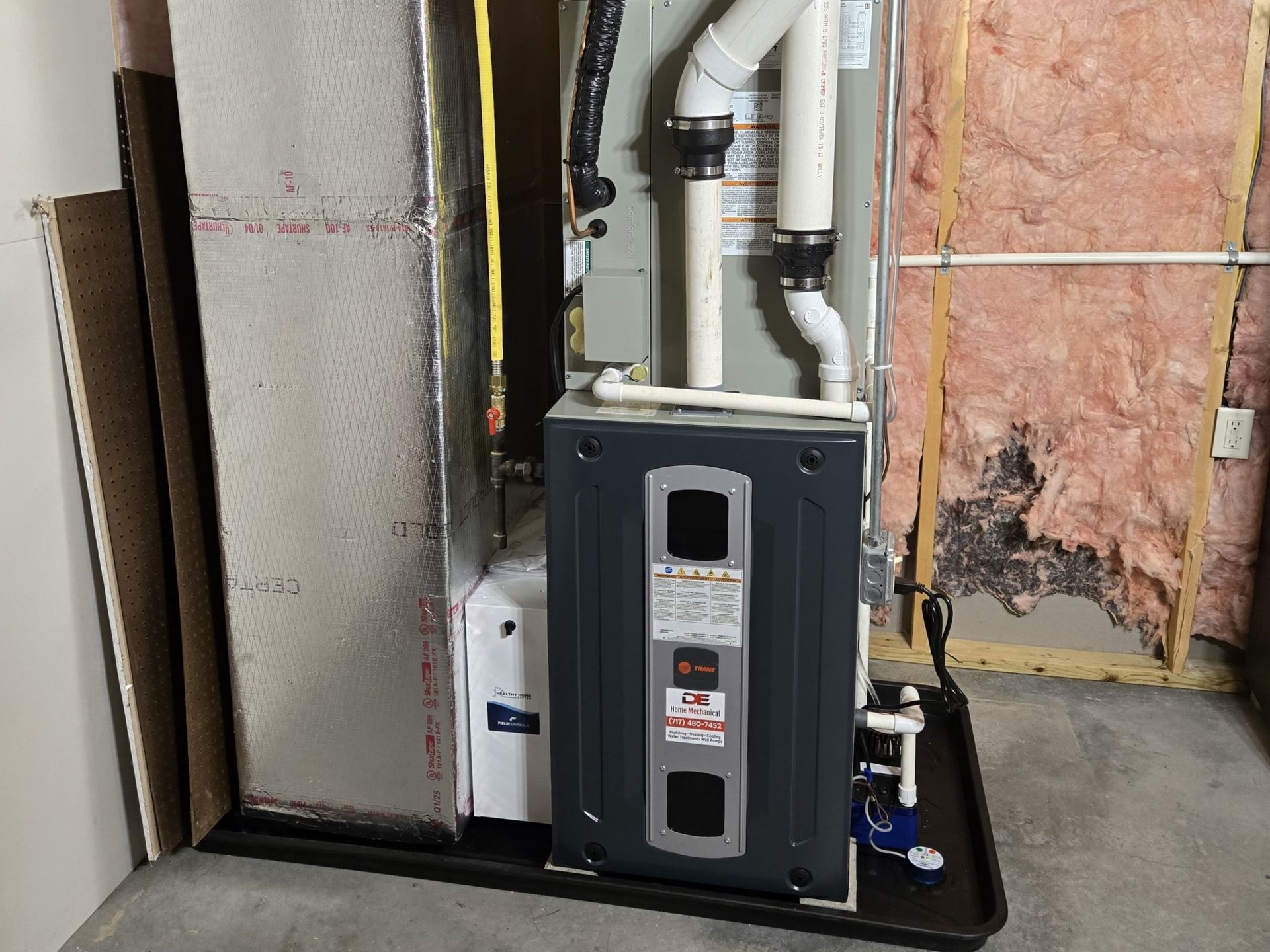Furnace unit in a utility room. unit with white pipes and a drain pan, set against a concrete floor and insulation.