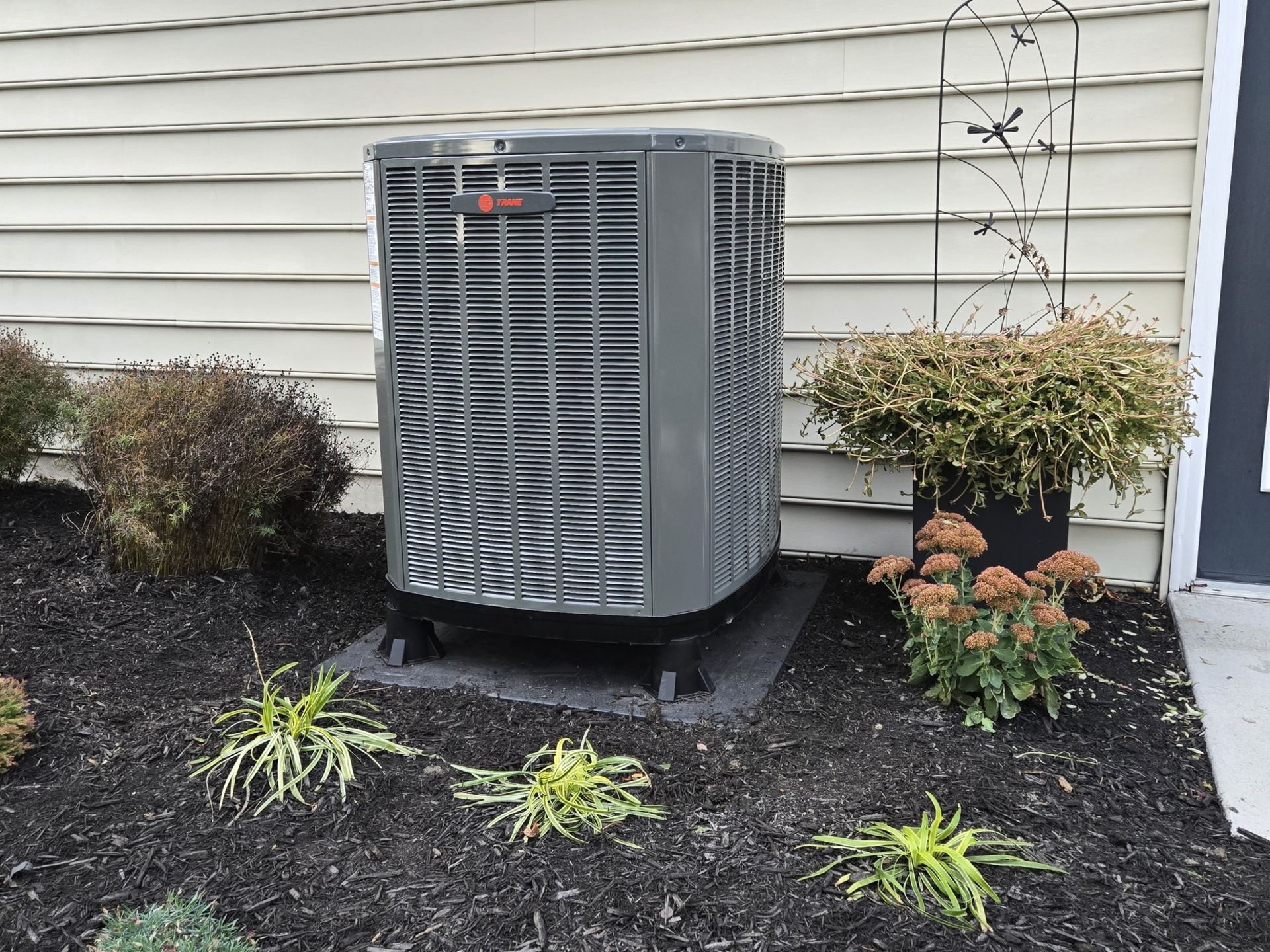 Outdoor HVAC unit on a black base, surrounded by mulch and greenery, near a building's siding.