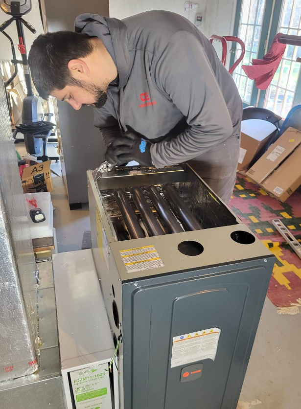 A man in gray shirt is repairing a furnace, indoors. He is wearing gloves and looking inside.