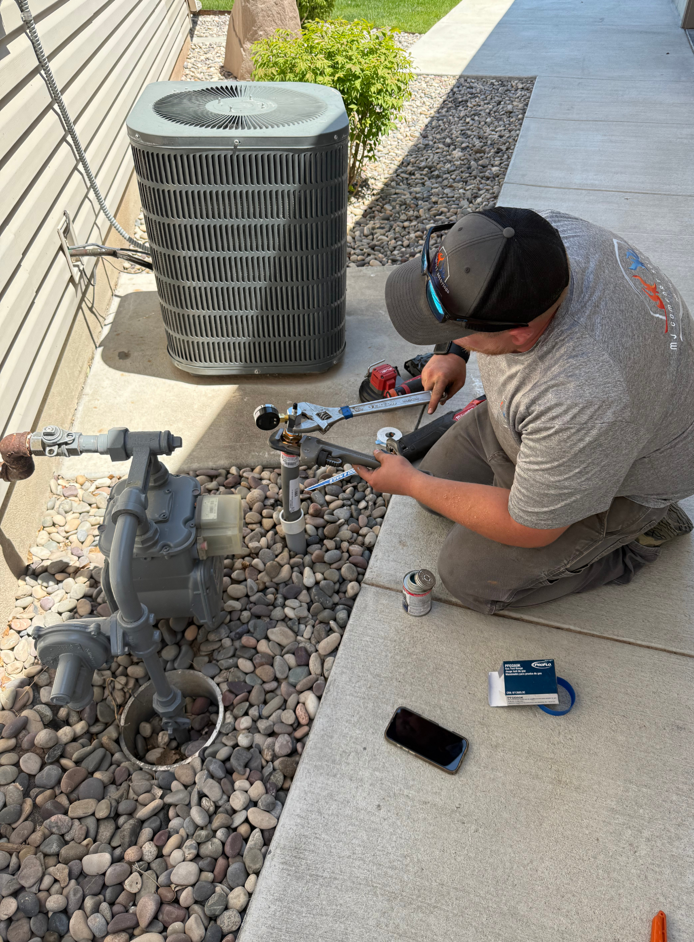 Man kneeling, working on a gas meter outside, with AC unit and house siding in the background.
