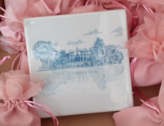 Piastrella quadrata con disegno paesaggistico blu, circondata da sacchetti regalo in organza rosa.