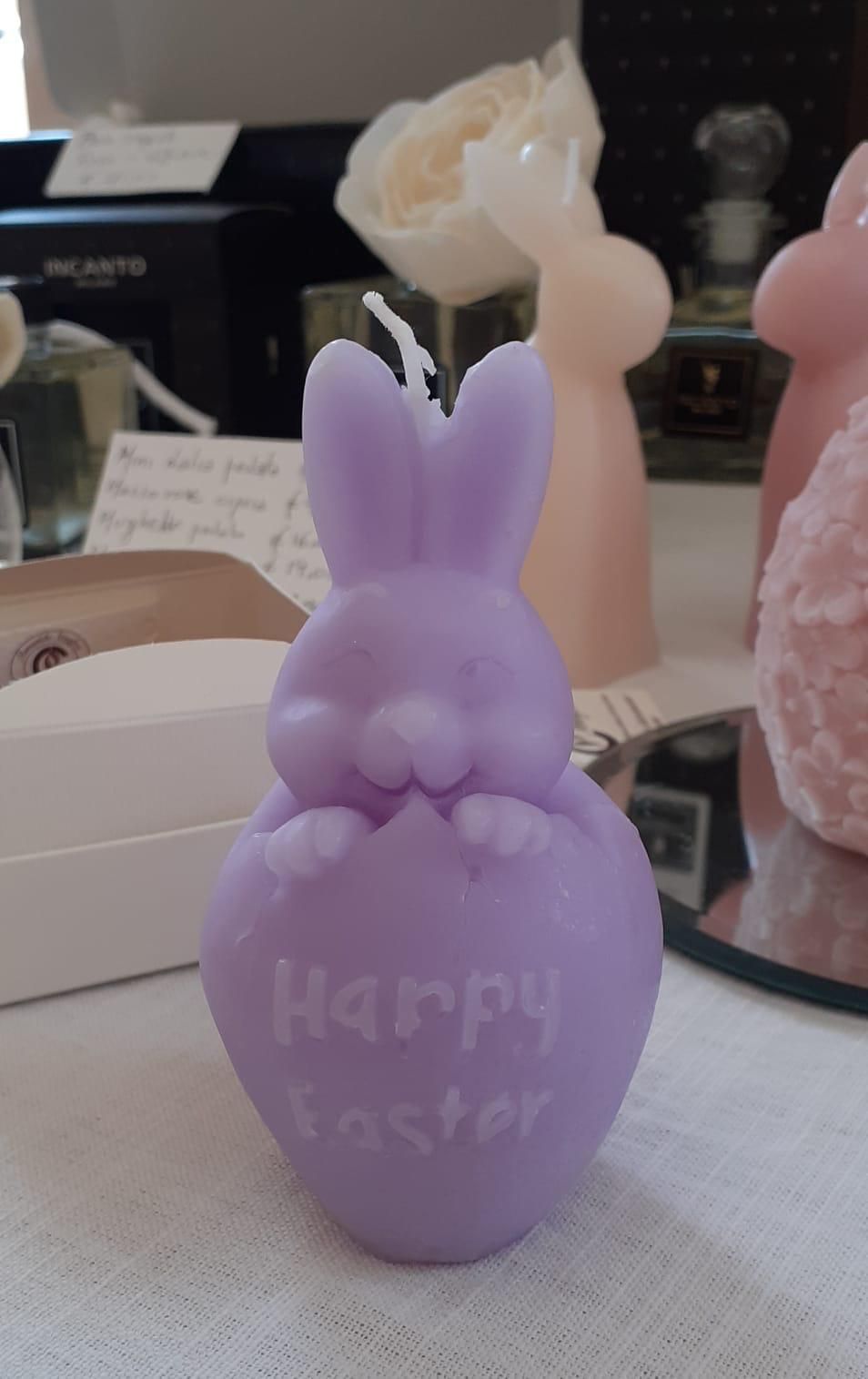 Una candela a forma di coniglietto color lavanda che esce da un uovo con la scritta