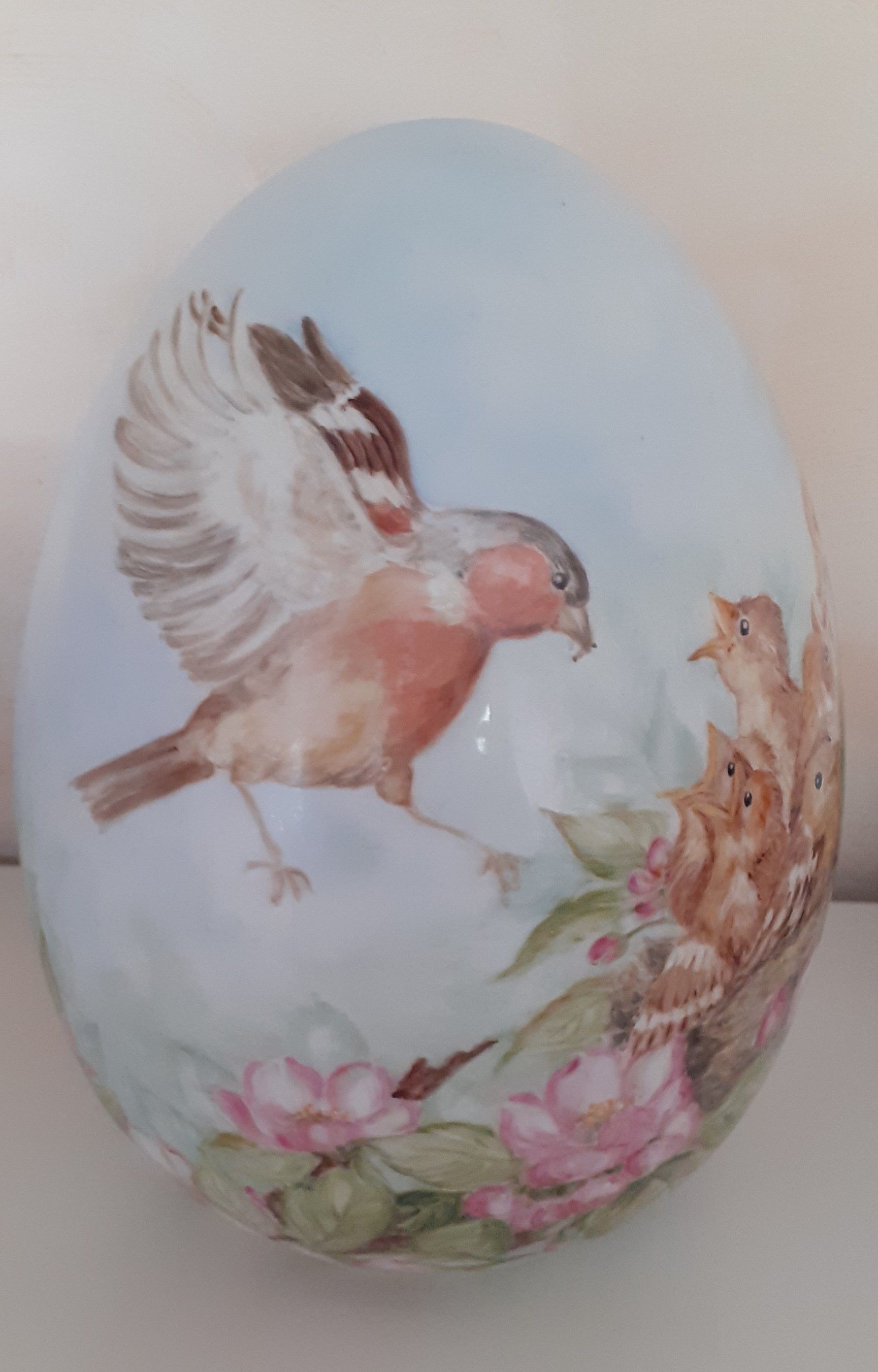 Uovo dipinto con un uccellino che nutre i suoi nidiacei; fiori rosa e cielo azzurro.