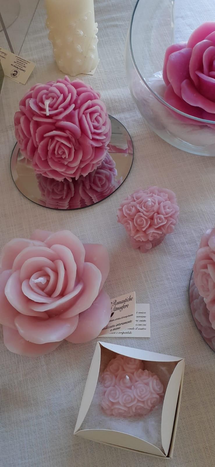 Candele rosa a forma di rosa di varie dimensioni su una superficie bianca, alcune in ciotole e scatole di vetro.