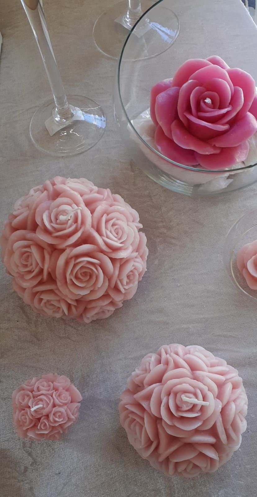 Candele rosa a forma di rosa e una sfera floreale su una tovaglia bianca, vicino a bicchieri di vino vuoti.