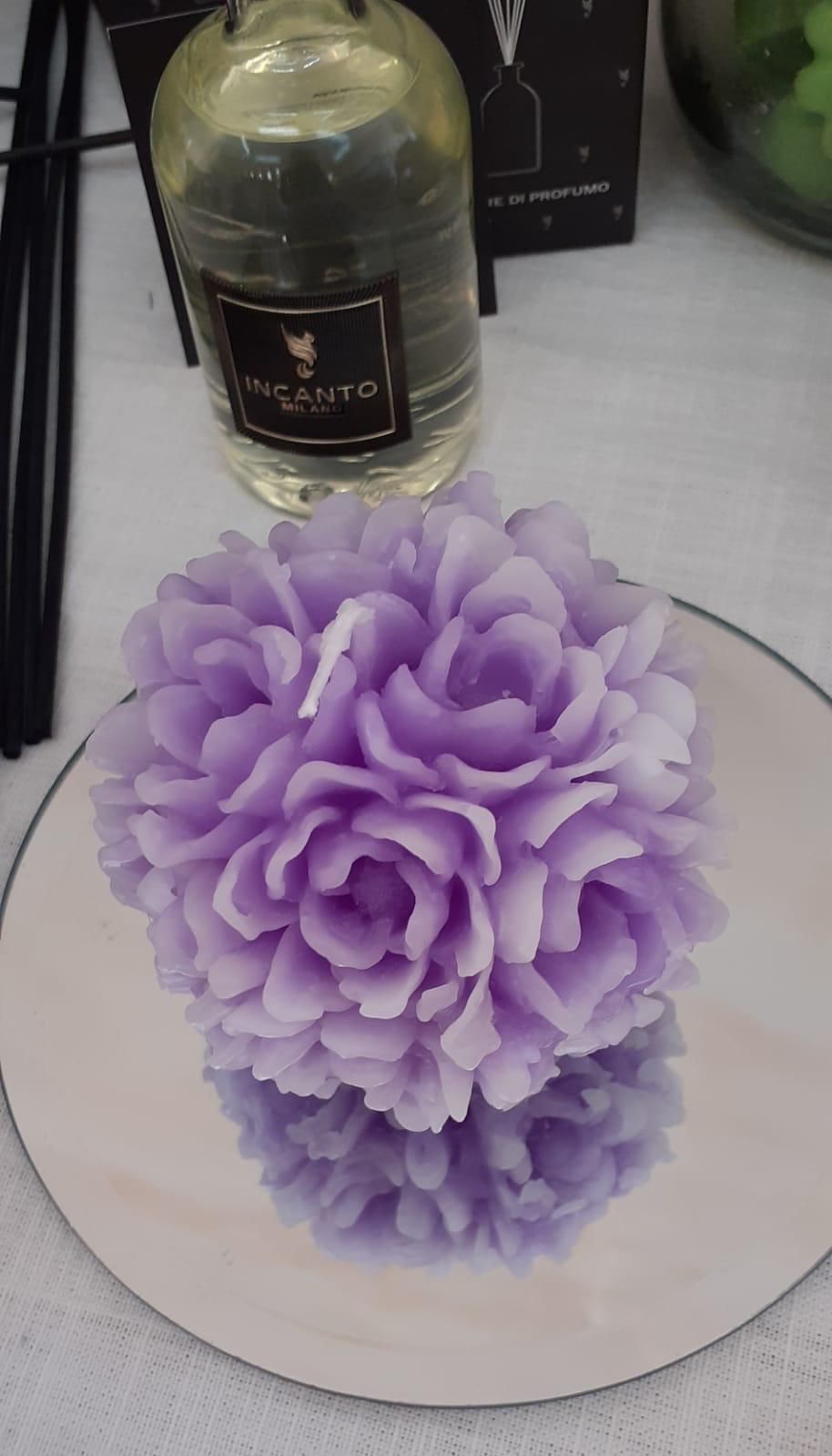 Candela viola a forma di fiore su una superficie riflettente, accanto a una bottiglia di profumo e a dei bastoncini.