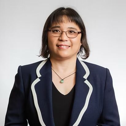 Portrait of a smiling person wearing glasses and a navy blazer with white trim on a light gray background
