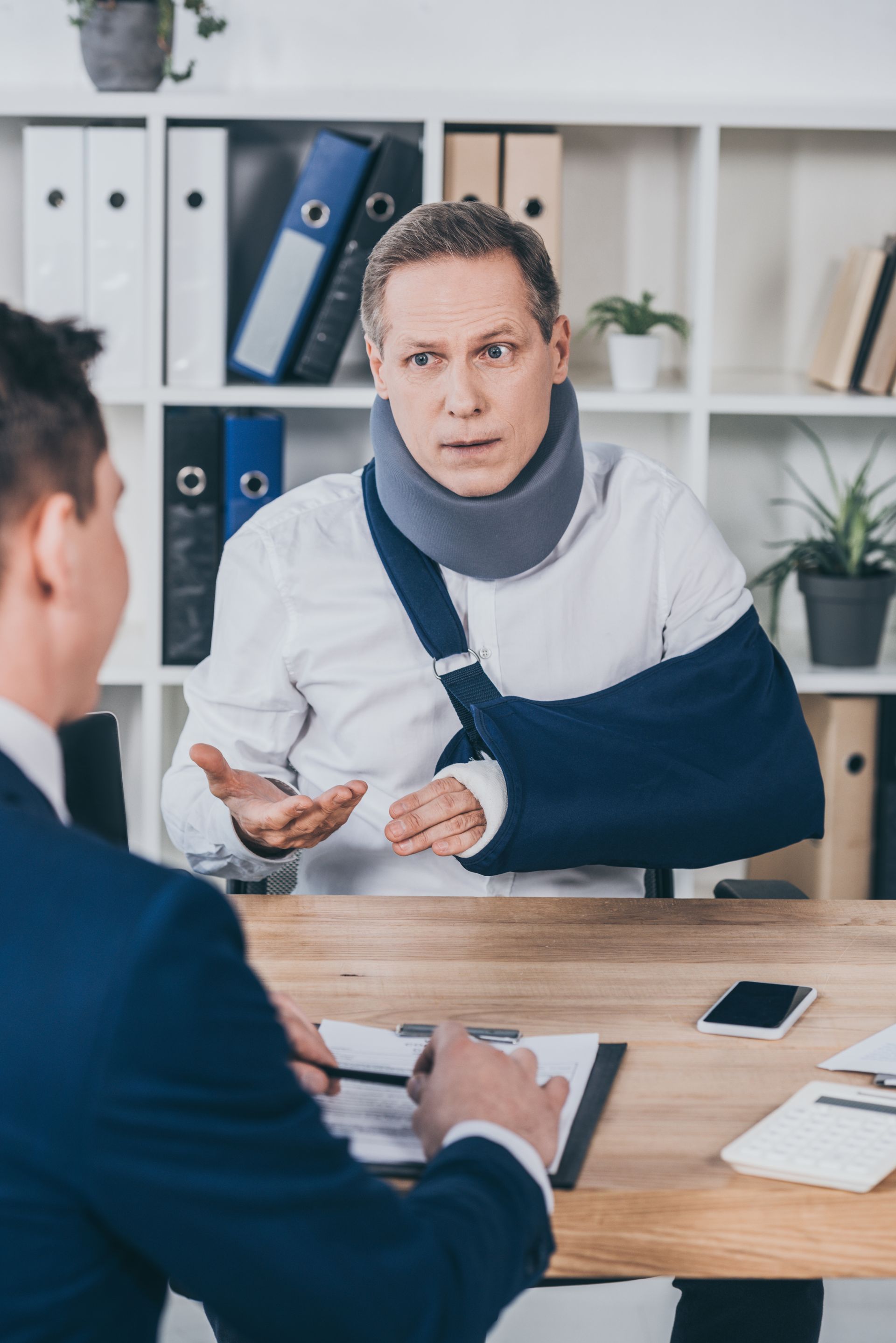 Person with arm in sling speaking during a meeting at a desk with notebooks and a phone.