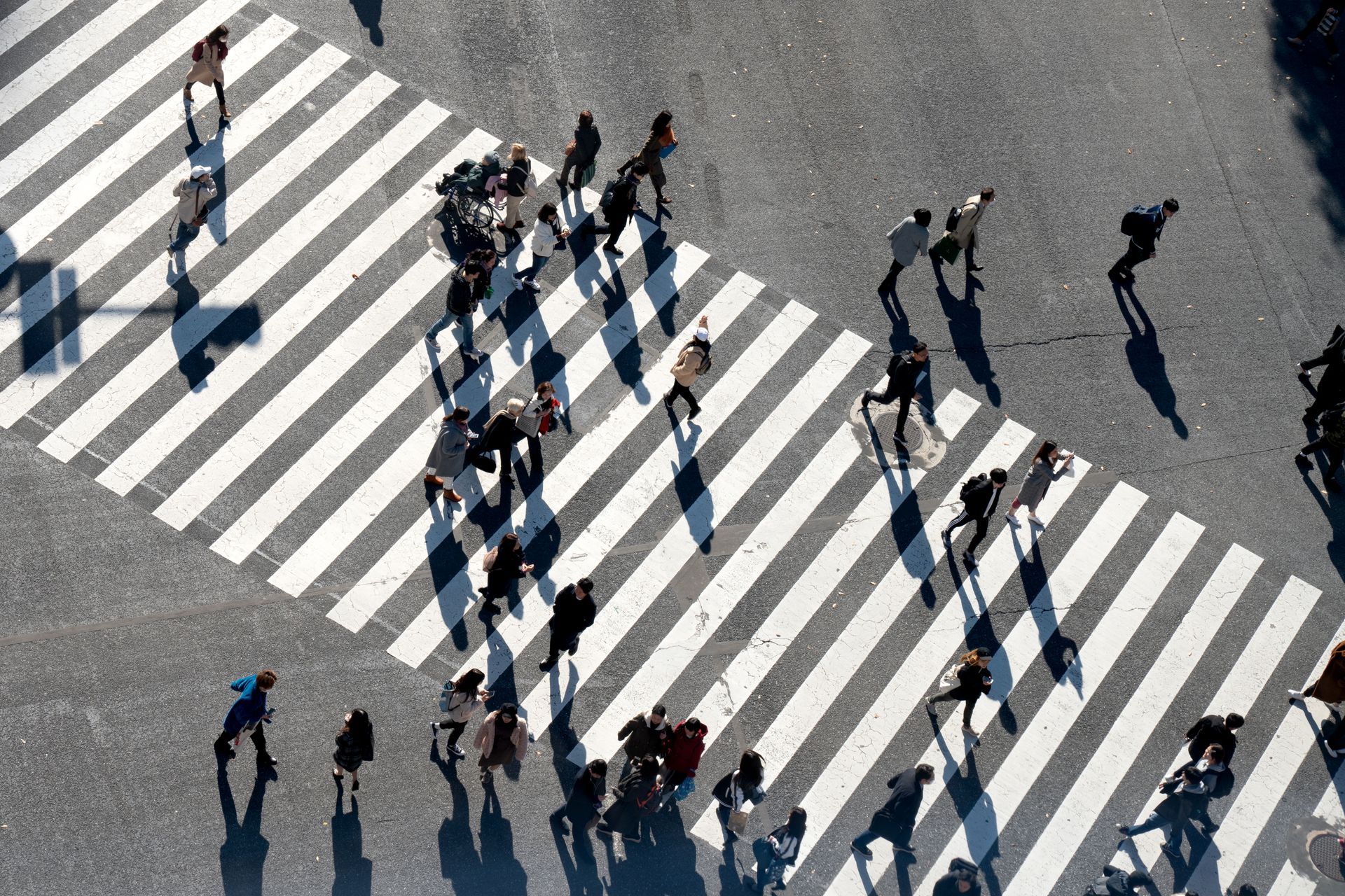 Pedestrians crossing a wide striped crosswalk from above, with long shadows on gray pavement