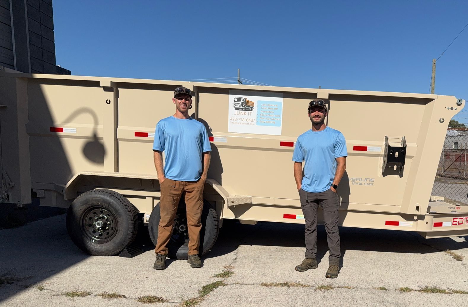 Two men in blue shirts stand by a tan trailer on a sunny day.