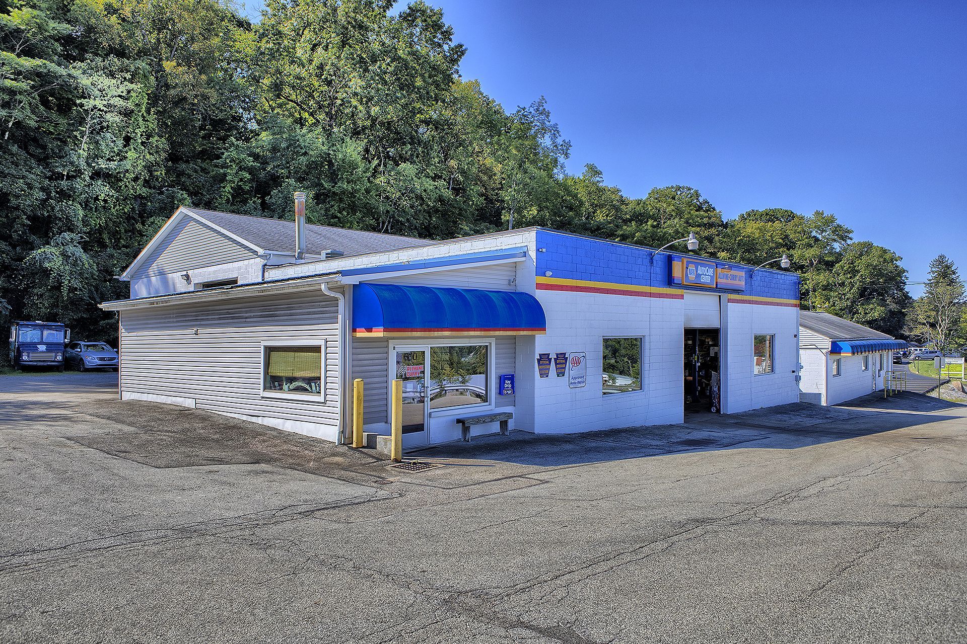 A single-story commercial building with white walls, a blue striped awning, and a gravel lot against a backdrop of trees. | Allwine Curry Inc