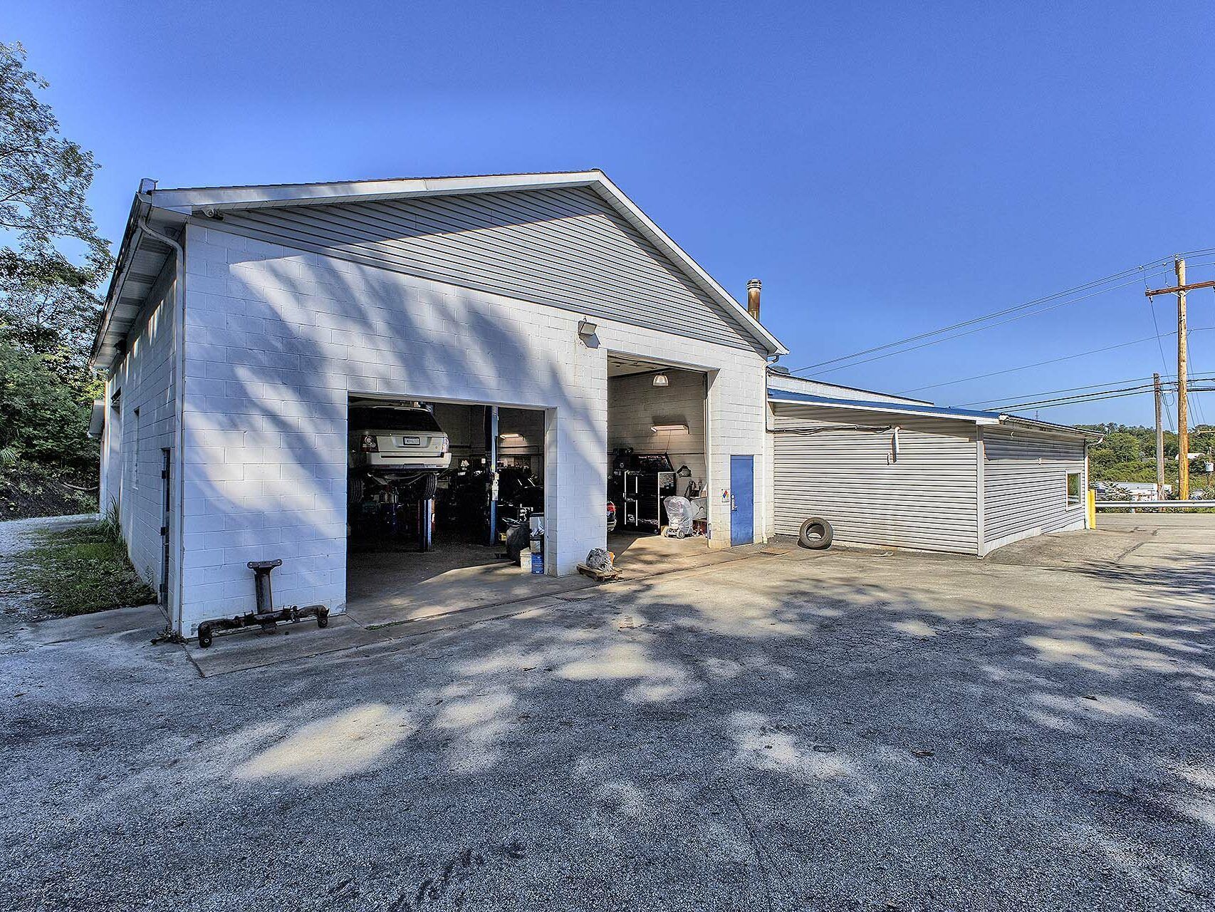 A white, single-story garage with two open bays, a gravel lot in front, and a side extension with corrugated metal walls. | Allwine Curry Inc
