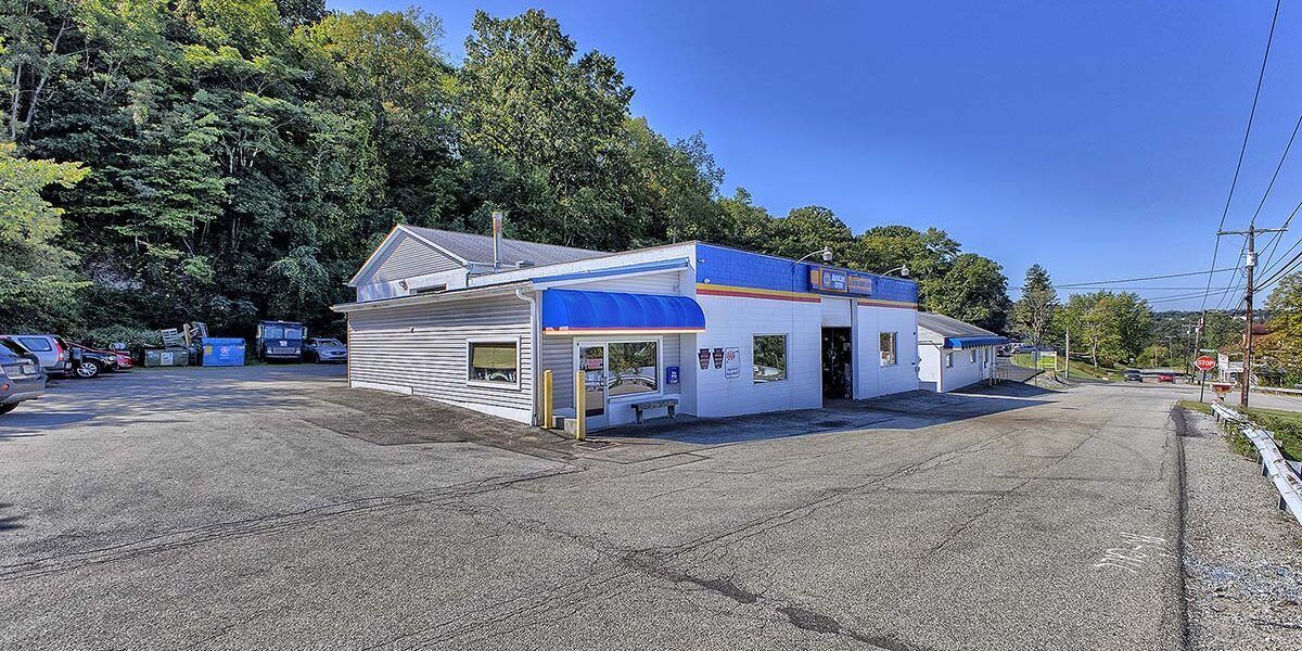 A white, one-story commercial building with blue awnings sits on a gravel lot next to a wooded hillside under a blue sky. | Allwine Curry Inc