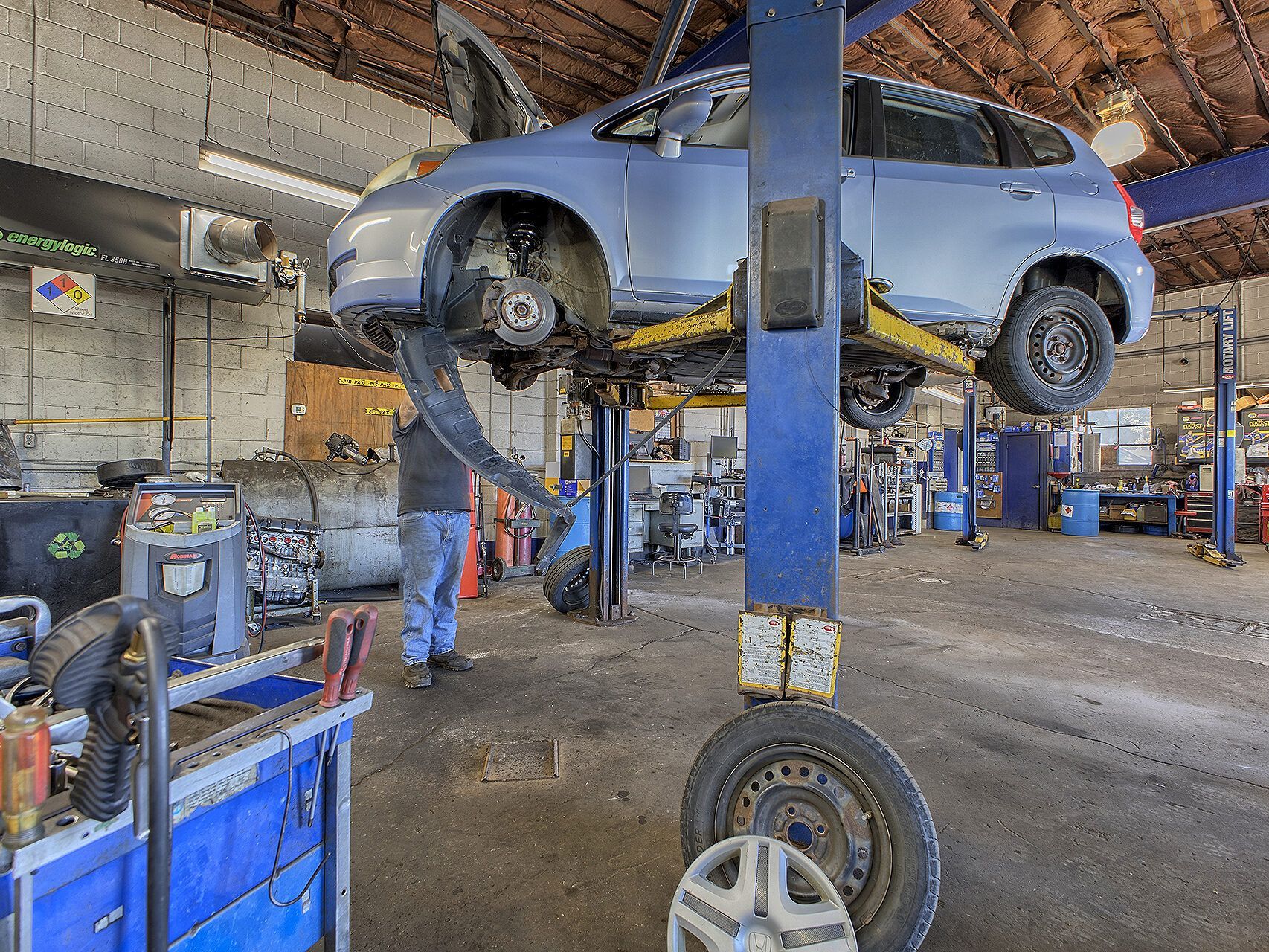 A light blue hatchback lifted on a garage hoist, with a mechanic working and a detached tire in the foreground. | Allwine Curry Inc