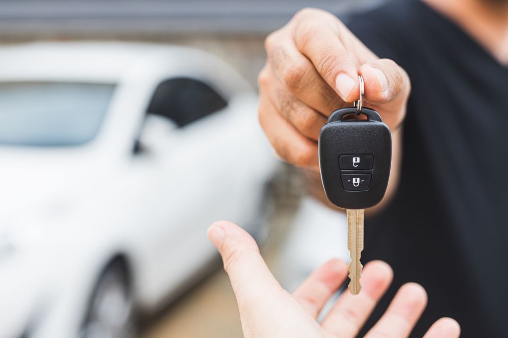 A person handing a car key to another person, with a blurred white car in the background. | Allwine Curry Inc