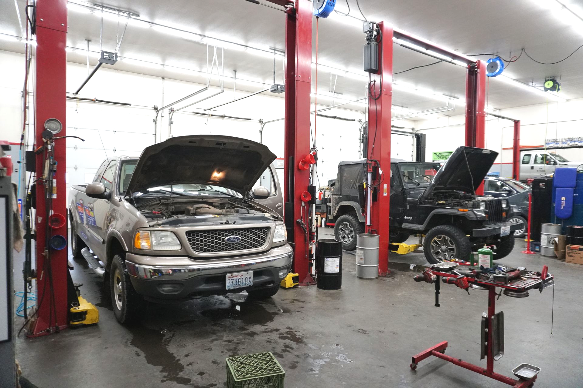 A repair shop with two vehicles on lifts. One is a pickup truck, the other a Jeep, both with open hoods.