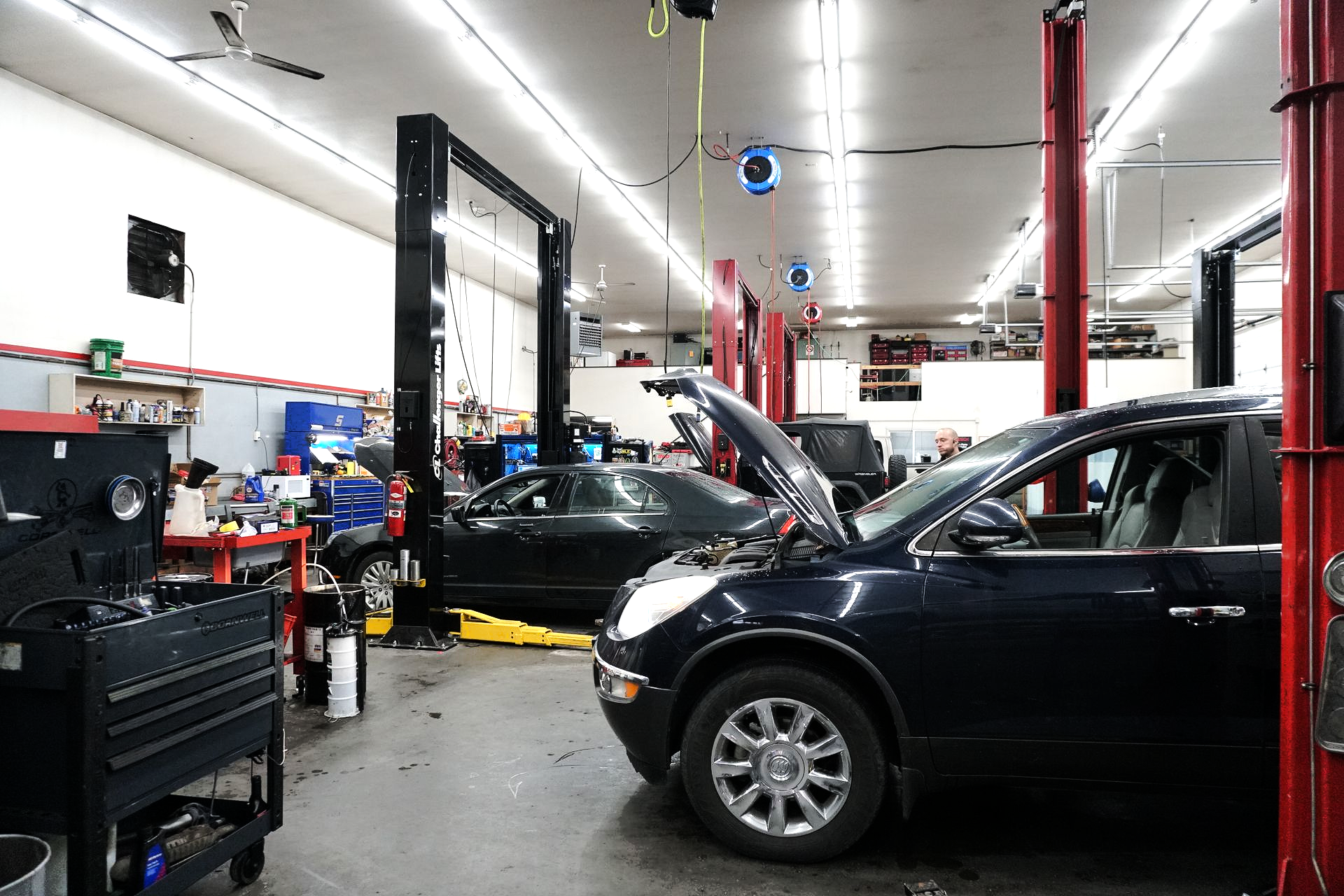 Car repair shop, two cars on lifts with hoods open, tools and equipment visible, fluorescent lighting.