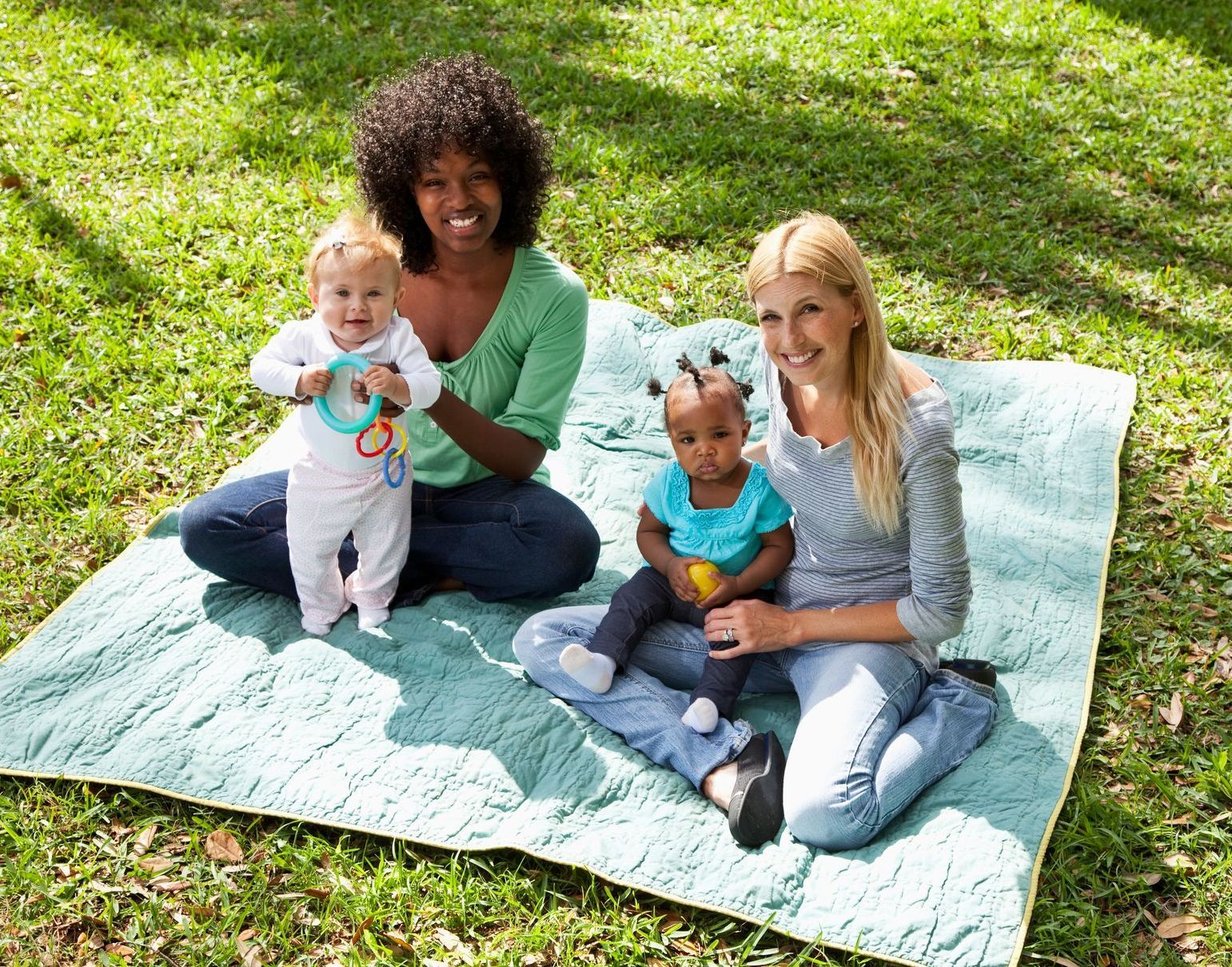 Two moms and their babies sitting on a blanket in the park.