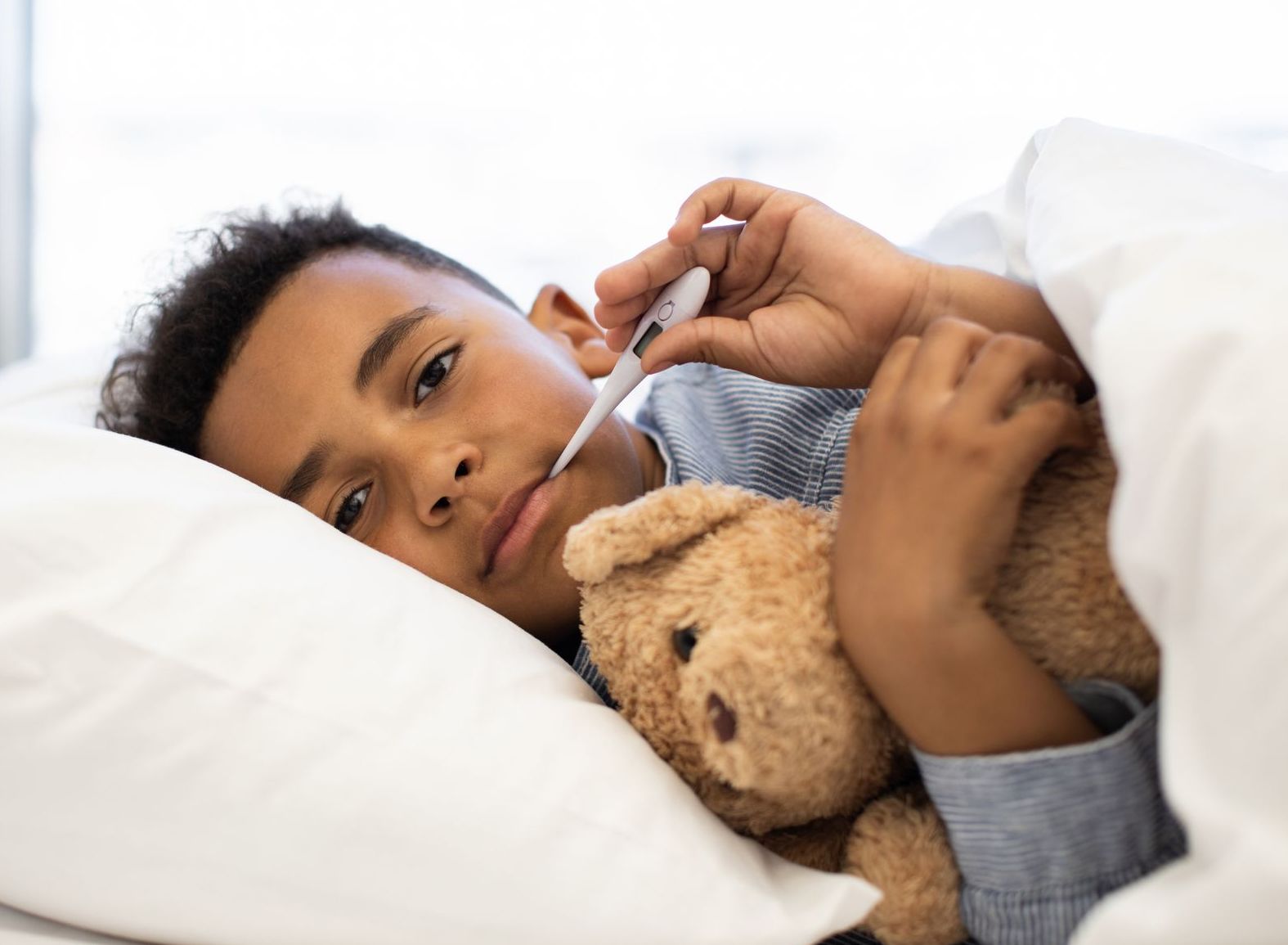 A boy in bed with a teddy bear and a thermometer in his mouth.