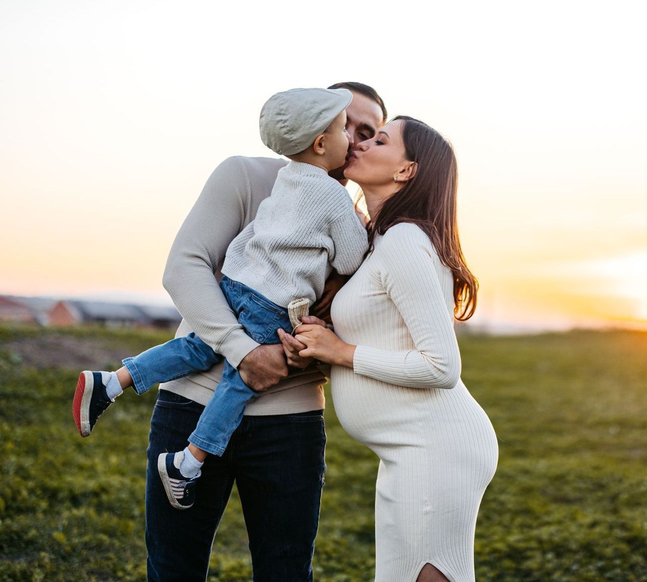 A pregnant mother and father hold their son with their faces close together. There is a sunset and grassy area in the background.