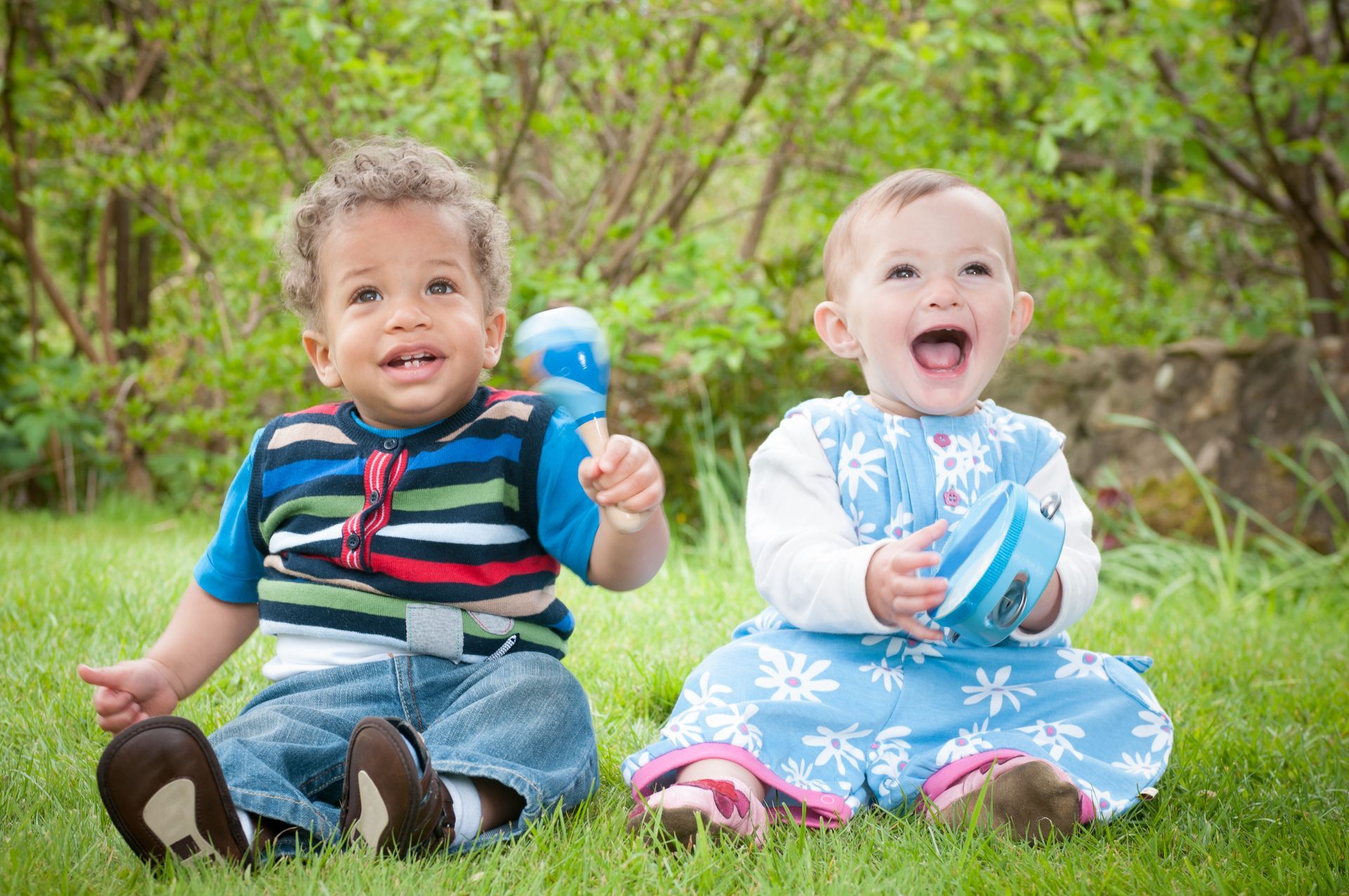 Two babies sitting on the grass in a park. They are both smiling. 