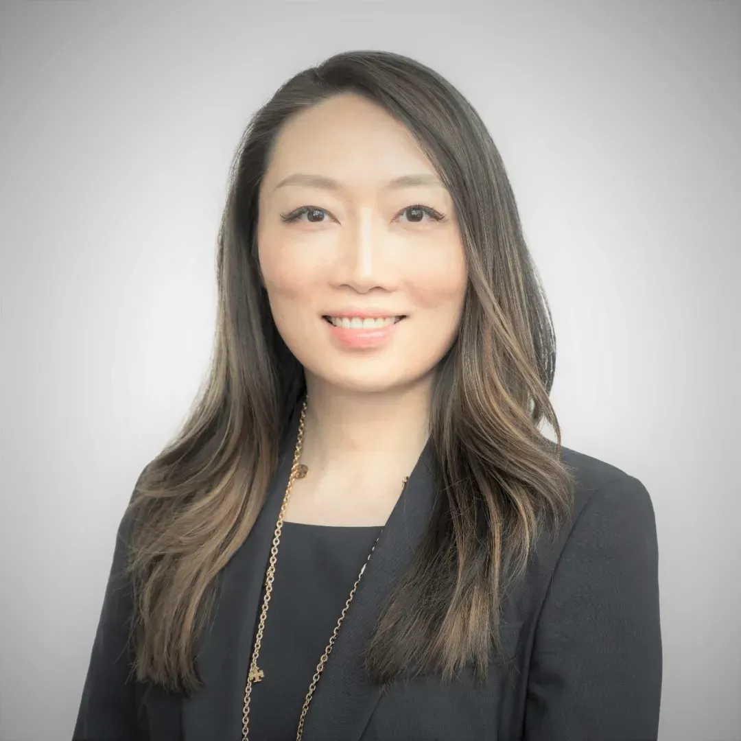 Woman with long brown hair, wearing a black blazer and a gold necklace, smiling at the camera.