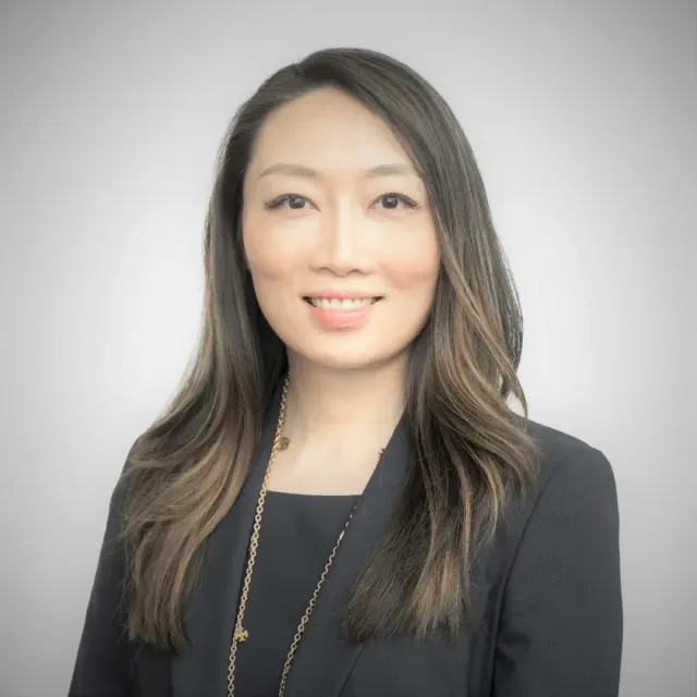 Woman with long brown hair, wearing a black blazer and a gold necklace, smiling at the camera.