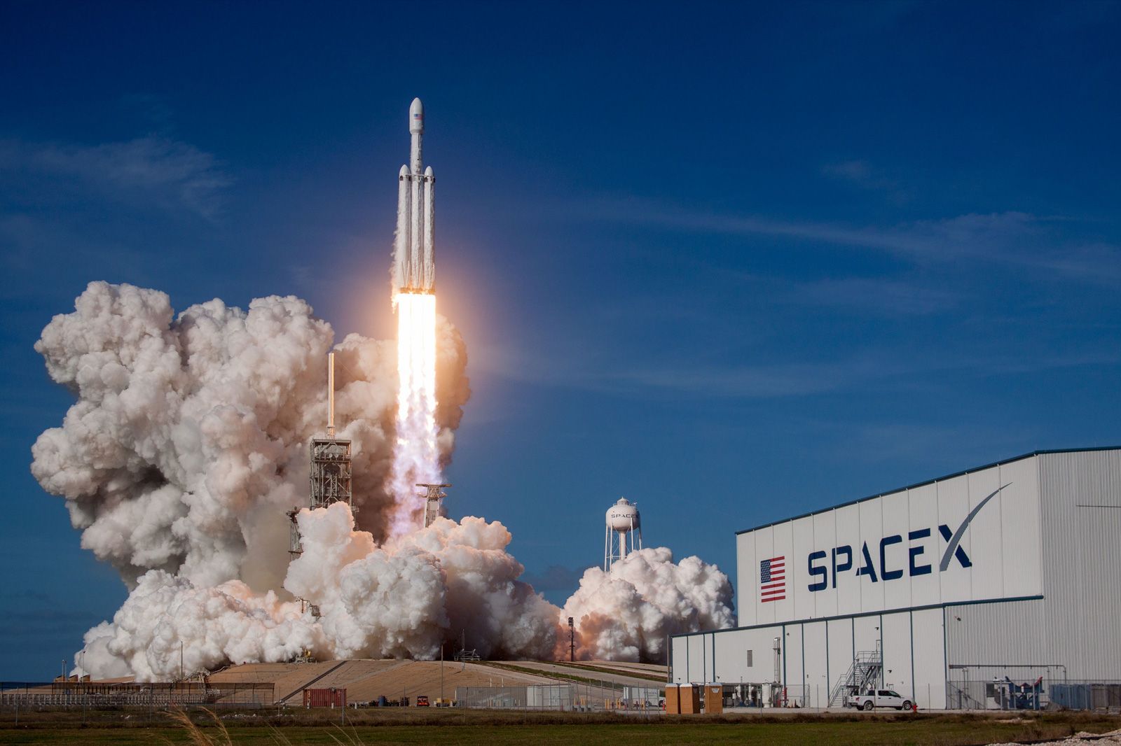 SpaceX Falcon Heavy rocket launching, white smoke, bright orange flames, blue sky, SpaceX building visible.