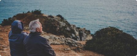 Two people in jackets sit on a rocky cliff, looking out over the ocean.