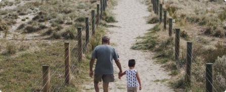 Two people walk hand in hand along a sandy path through grassy dunes