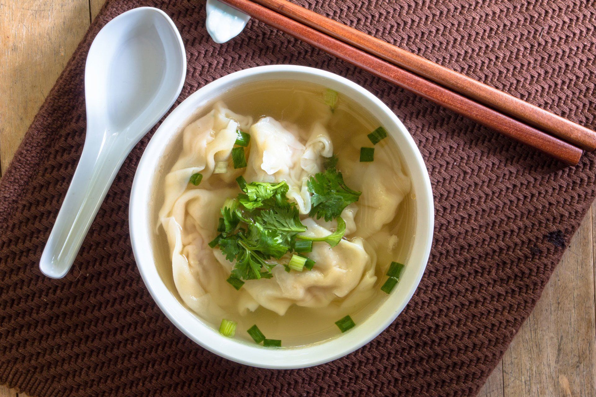 Bowl of dumpling soup topped with herbs, beside a white spoon and chopsticks on a brown mat.