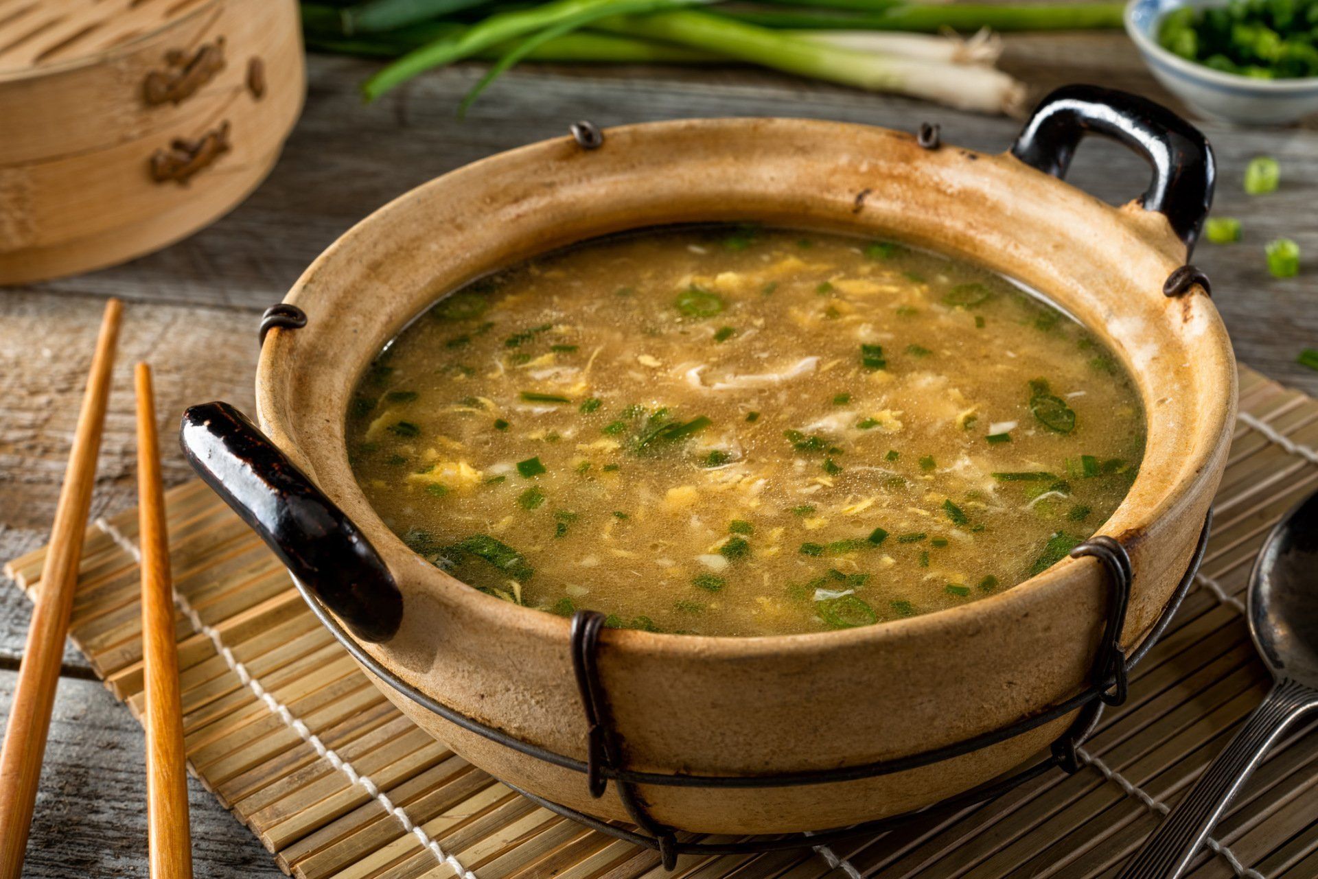 Clay pot of green soup with chopped herbs on a bamboo mat, with chopsticks beside it.