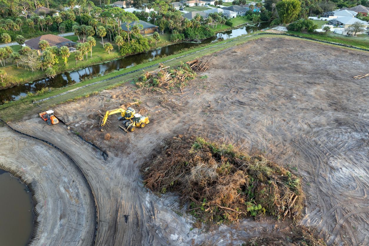 Aerial view of a cleared dirt lot with construction machinery and a brush pile beside a residential area