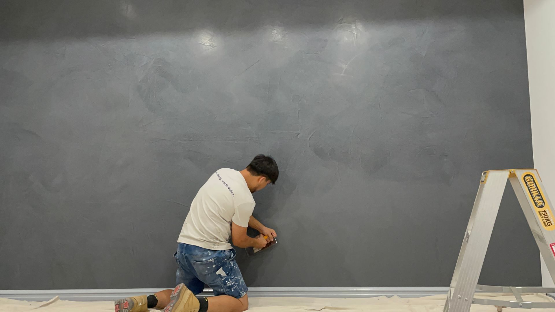 Person kneeling, applying something to a dark gray textured wall. A step ladder is beside them.