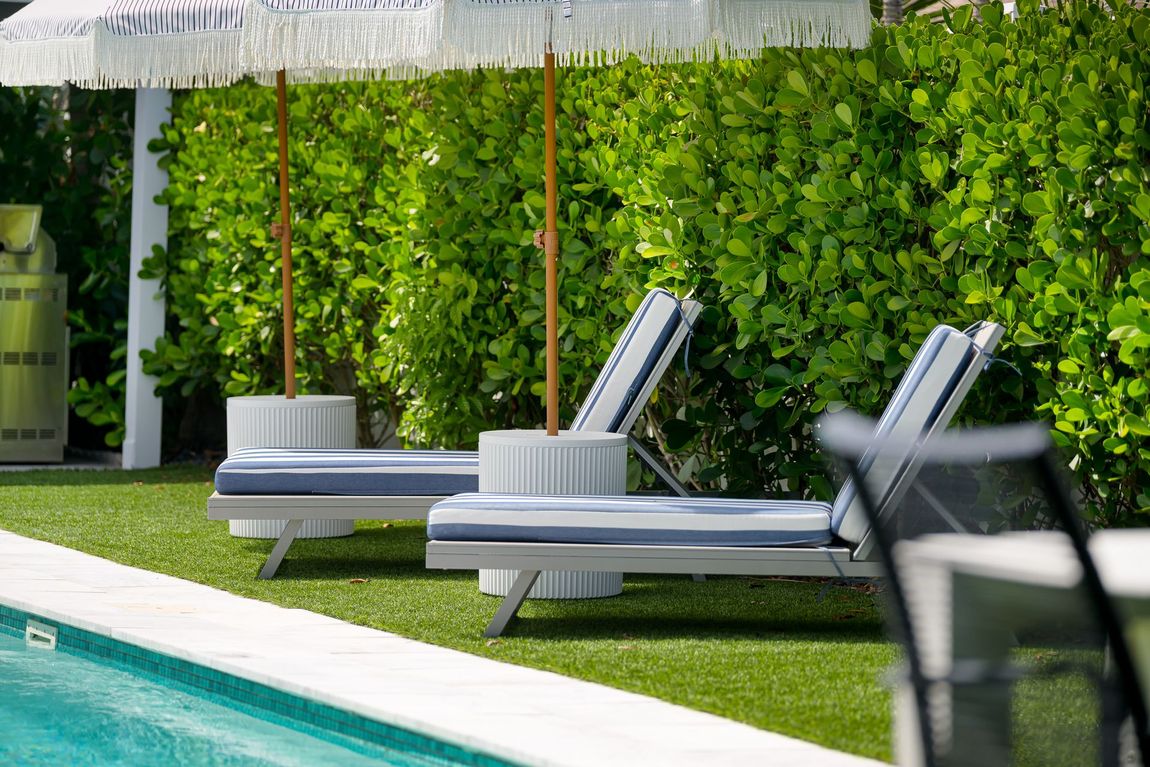 Poolside lounge chairs beside a green hedge and turquoise swimming pool under a white umbrella.